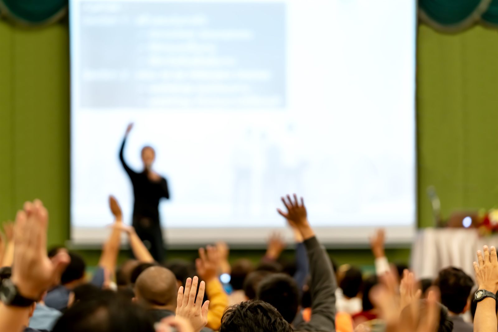 Presenter in front of a projection screen as attendees raise their hands